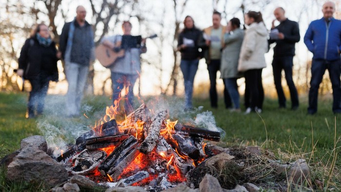Eine Gruppe versammelt sich um ein lebhaftes Lagerfeuer im Freien auf einer grasbewachsenen Wiese und genießt die Gesellschaft der anderen.