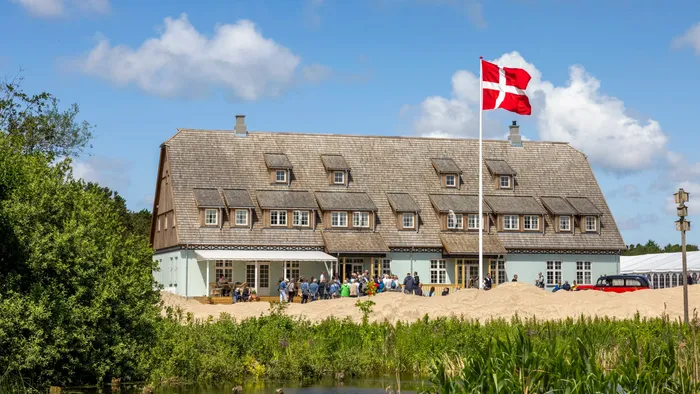 Et malerisk bygning med et dansk flag, omgivet af grønne områder og en dam, der er vært for en udendørs sammenkomst.