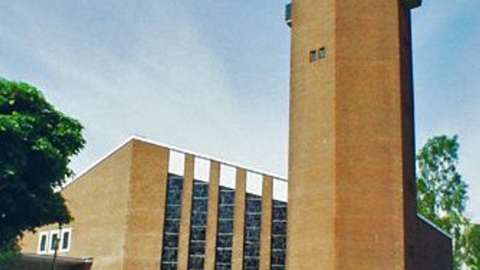 Das Bild zeigt einen hohen Ziegelbau mit einem Turm und umgeben von Grün.