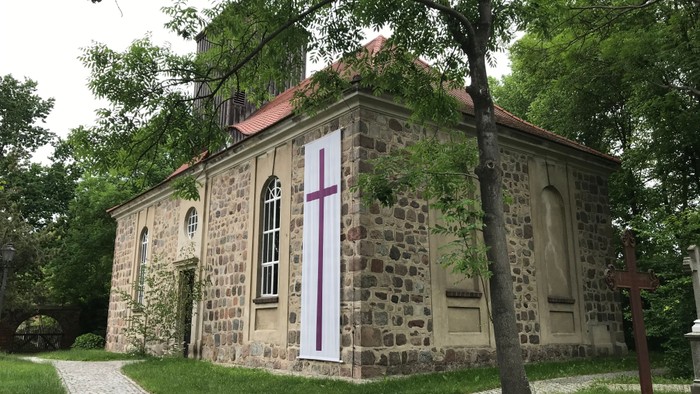 Das Bild zeigt eine steinerne Kirche mit einem Kreuz an der Fassade, umgeben von Bäumen und einem Kopfsteinpflasterweg.