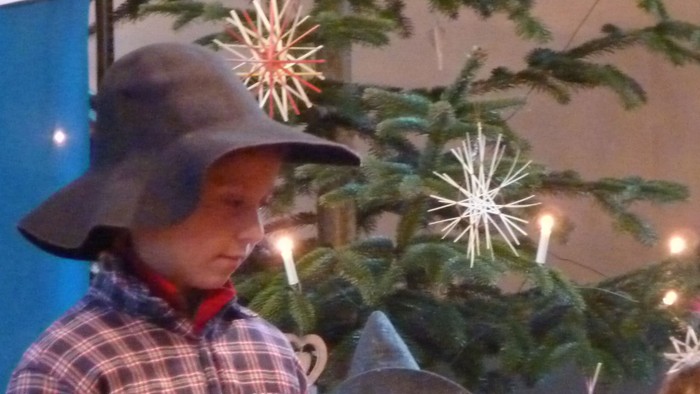 Kinder in Weihnachtskleidung, eines hält ein Mikrofon, mit einem geschmückten Baum im Hintergrund.
