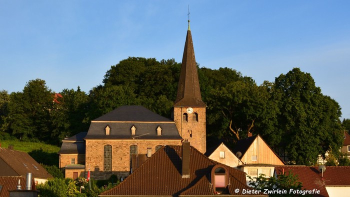 Das Bild zeigt die Evangelische Kirche Herbede mit einigen Häusern vor einem blauen Himmel.