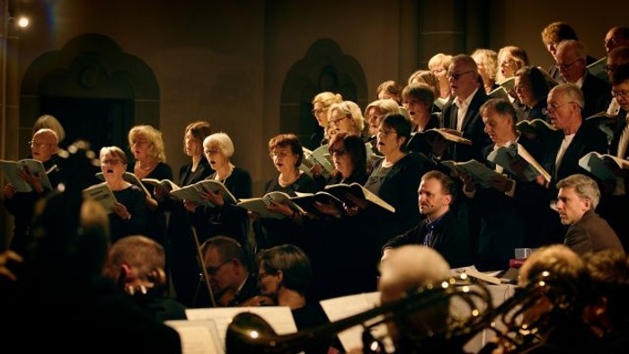 Ein Chor und ein Orchester treten gemeinsam in einem großen Konzertsaal auf und harmonisieren mit Notenblättern.
