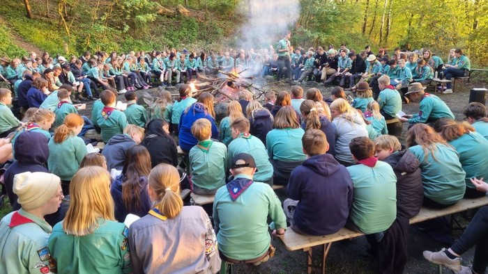 Große Gruppe von Menschen in einheitlichen T-Shirts, die sich im Freien um ein Lagerfeuer in einem bewaldeten Gebiet versammelt haben.