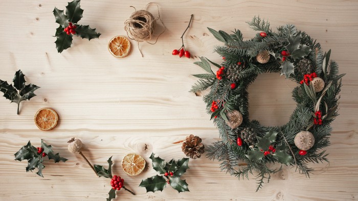 En julekrans med grønne nåler og røde bær, omgivet af tørrede apelsiner og granatæbler på et hvidt træ.