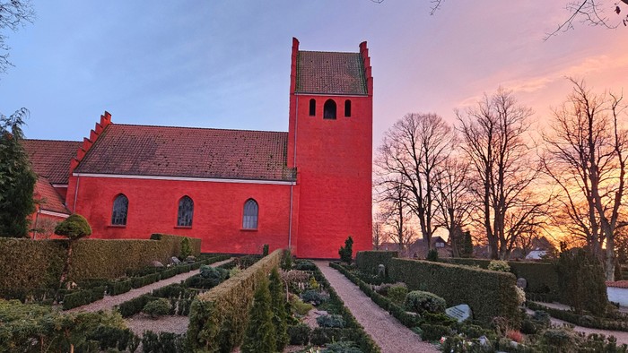 Livligt rødt kirkebygg med omhyggeligt anlagt have under en farverig solnedgangshimmel.