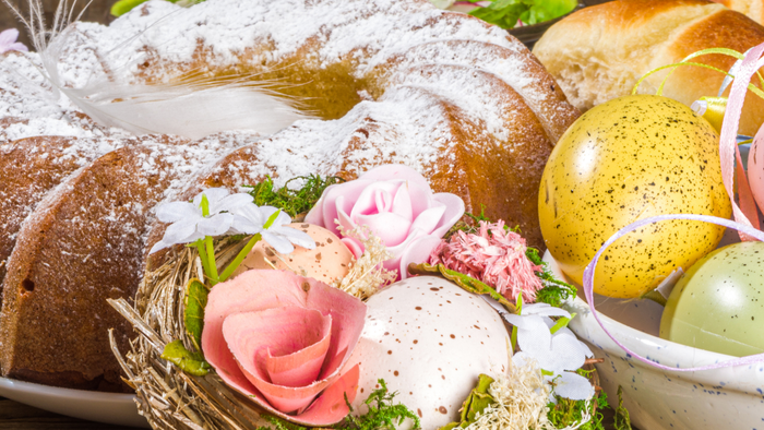 Bunte Ostereier, Gebäck und verziertes Brot in einem festlichen Korb mit Blumen und Grünzeug.