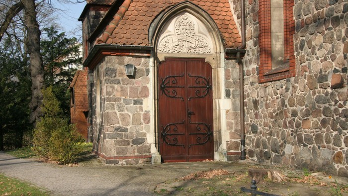 Das Bild zeigt den Eingang einer Kirche mit einem hölzernen Tor und einem steinernen Gebäude.