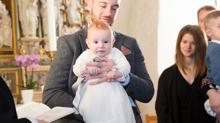 En mand holder et spæd barn i en kirke, mens en præst læser fra en bog. En kvinde og et andet barn står ved siden af dem.