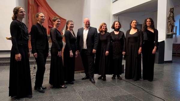 Eine Gruppe von neun Personen in formeller Kleidung, die drinnen gemeinsam posieren, wahrscheinlich ein Chor oder ein Ensemble für eine Aufführung.