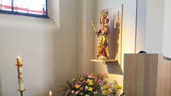 Auf einem Altar stehen eine Statue, Blumen und Kerzen vor einem Fenster