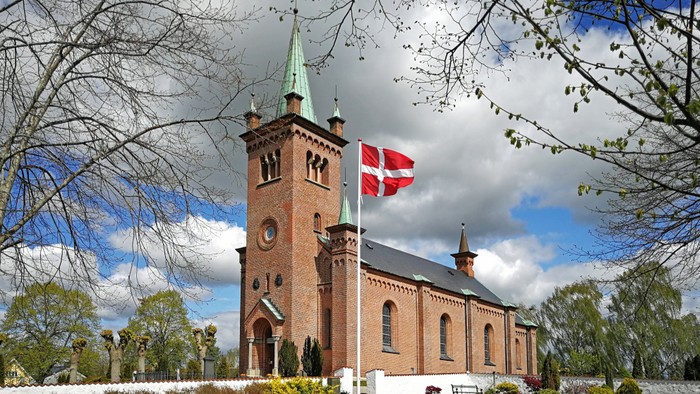 En malerisk rødstenskirke med et dansk flag, der vajer, omgivet af blomstrende træer og en fredfyldt kirkegård.