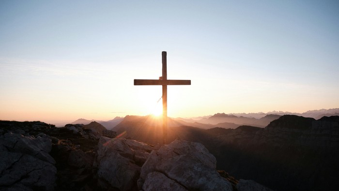 Eine Silhouette eines Kreuzes ragt markant über schroffen Felsen bei Sonnenaufgang über fernen Bergen empor.