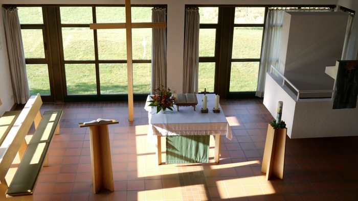 Heller Kapelleninnenraum mit hölzernen Kirchenbänken, zentralem Altar und natürlichem Licht, das durch große Fenster einfällt.