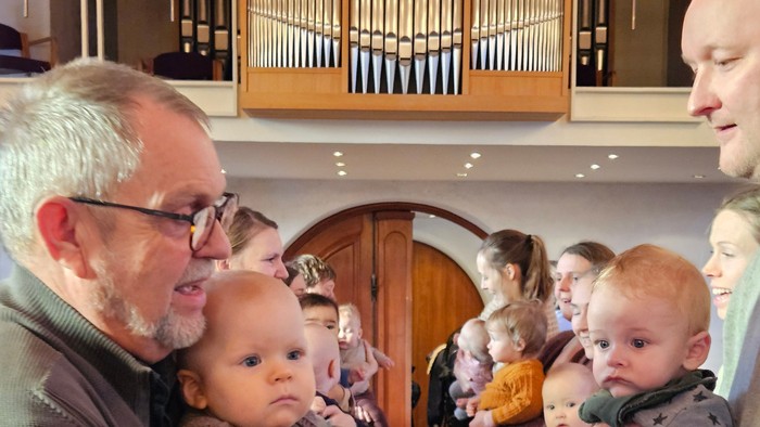 En mand holder en baby i en kirke, hvor andre er samlet, herunder børn og voksne, nær et orgel.