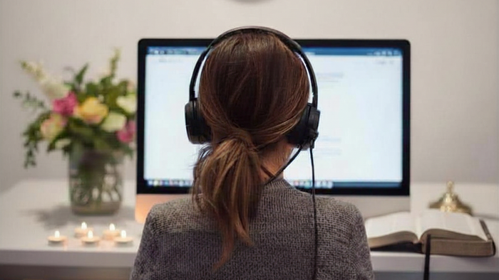 Eine Frau von hinten mit einem Headset. Sie sitzt vor dem Monitor. Auf der linken Seite sieht man Blumen und Kerzen auf dem Tisch. Auf der rechten Seite eine Bibel.