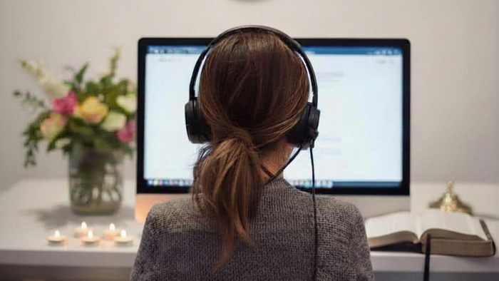 Eine Frau von hinten mit einem Headset. Sie sitzt vor dem Monitor. Auf der linken Seite sieht man Blumen und Kerzen auf dem Tisch. Auf der rechten Seite eine Bibel.