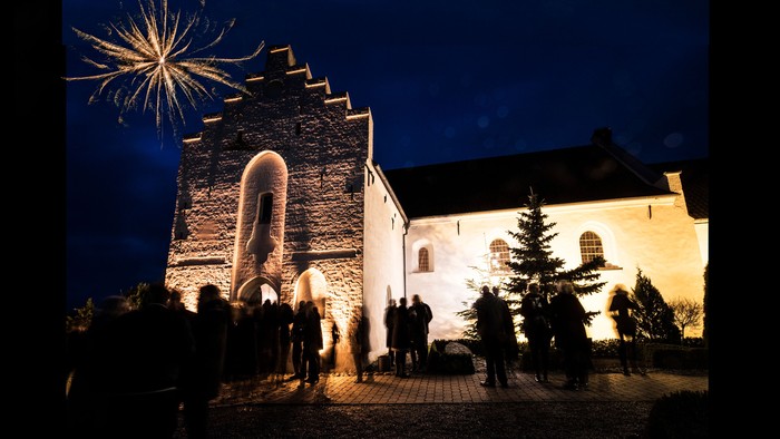 En mørk nattscene med en gruppe mennesker foran en belyset kirke og en tårn med fyrværkeri i himlen