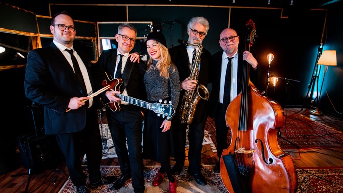 En jazzkvartet poser sammen på scenen med forskellige instrumenter, herunder guitar, saxofon og cello.