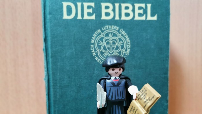 Eine Playmobil-Figur in Mönchsgewand hält ein Buch vor einem grünen Buch mit der Aufschrift 