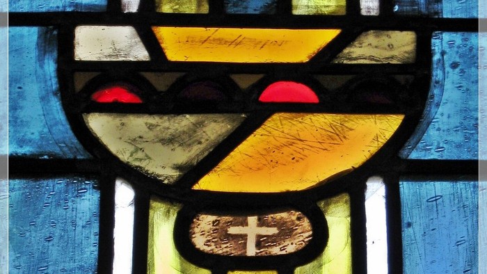 Das Bild zeigt ein buntes Kirchenfenster mit einem Kelch und Kreuz.