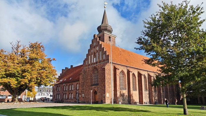 Historisk murstenskirke med højt spir omgivet af en frodig, grøn gårdsplads under en klar blå himmel.