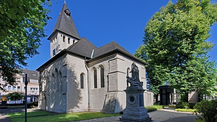 Kleine Kirche mit Turm und Statue davor, umgeben von Bäumen und Kopfsteinpflaster.