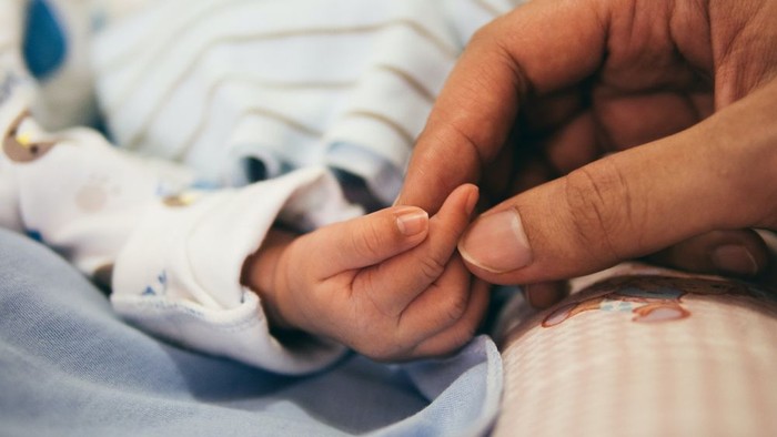 Eine Hand hält sanft die Finger eines Babys, das in eine blaue Decke gewickelt ist.
