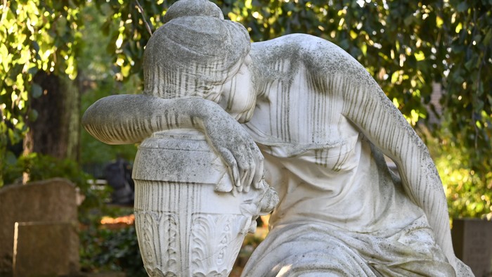 Eine Steinstatue einer Frau, die eine große, verzierte Vase in den Armen hält, in einer ruhigen Gartenlandschaft.
