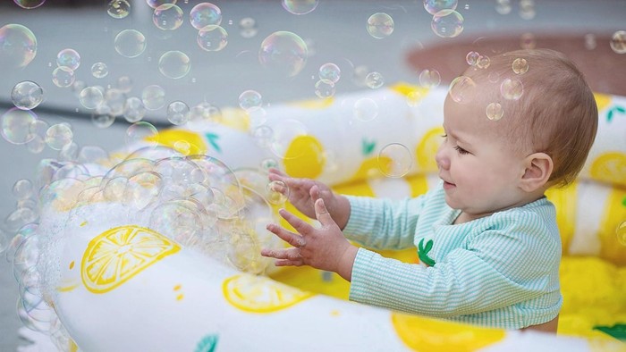 Ein Baby in einem aufblasbaren Pool mit Zitronenmuster streckt seine Hand aus, um die schwimmenden Blasen zu berühren, und lächelt vor Freude. Die Szene ist hell und