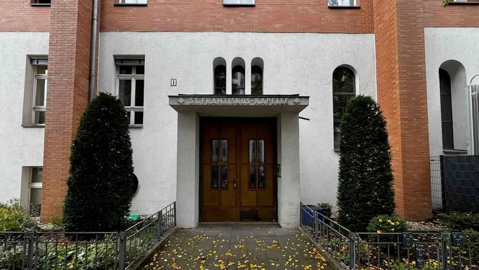 Das Bild zeigt den Eingang eines Gebäudes mit zwei Türen und Topfpflanzen.