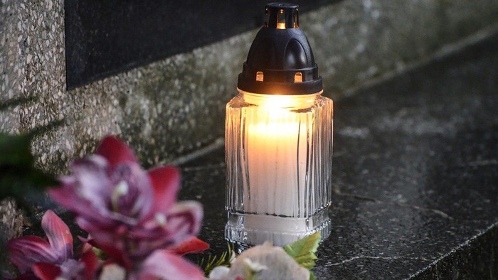 Eine brennende Kerze steht neben rosa Blumen auf einem grauen Stein.
