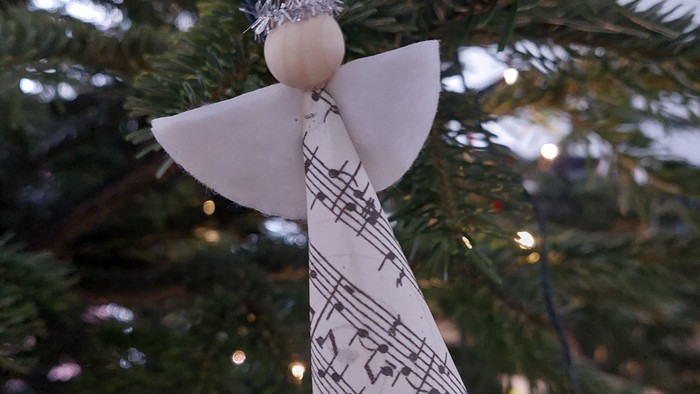 Håndlavet engelpynt med noder som vinger hængende på et pyntet juletræ.
