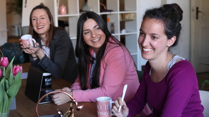 Drei junge Frauen sitzen an einem Tisch und trinken Kaffee, eine hält eine Tasse hoch.