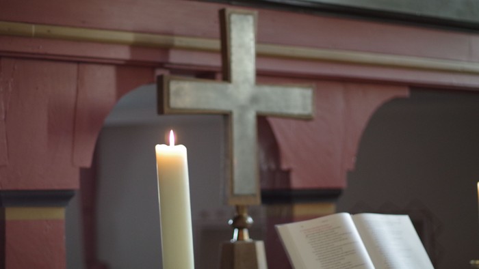 Eine brennende Kerze neben einem offenen Buch und einem Kreuz vor einem rosafarbenen Altarhintergrund.