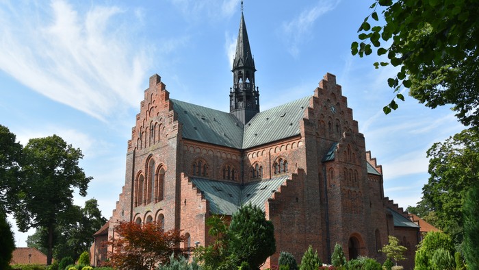 Historisk murstenskirke med højt spir omgivet af frodig grønne omgivelser under en klar blå himmel.