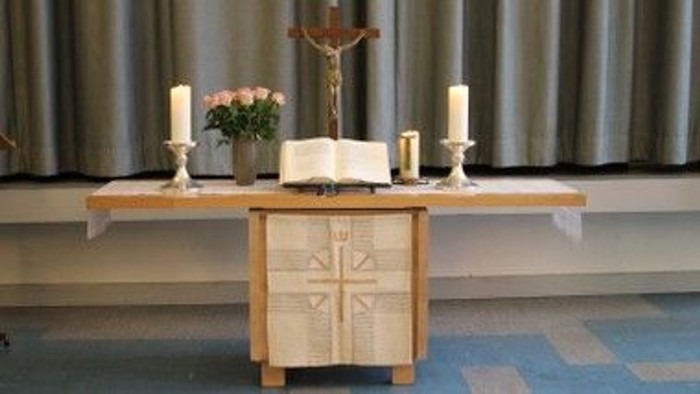Eine ruhige Altar-Anordnung mit einem Kreuz, Kerzen, Blumen und einem offenen Buch auf einem Holztisch.