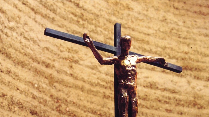 Ein Metallkreuz mit einem darauf befestigten Jesus-Figürchen vor einer Sandsteinmauer.