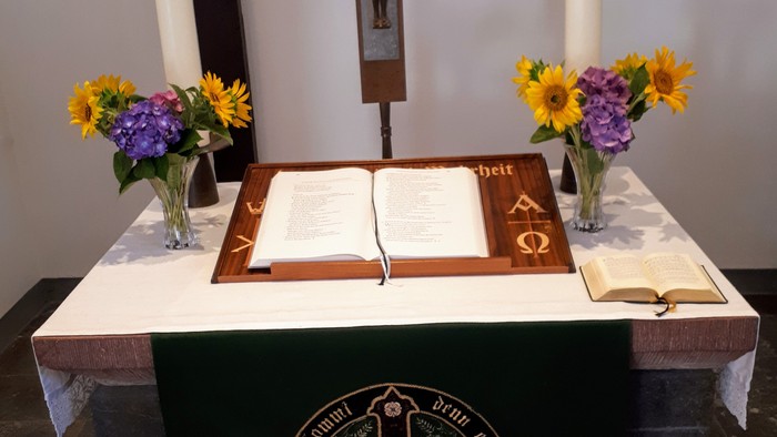 Religiöser Altar mit einer geöffneten Bibel, Kerzen, Blumen und einem Kruzifix auf einer verzierten Tischdecke.