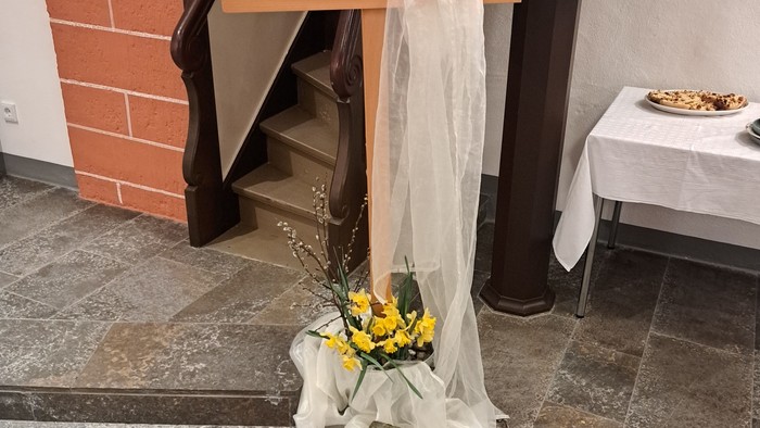 Eine mit weißem Stoff und Blumen drapierte Holzkreuz, das in der Nähe einer Treppe auf dem Boden platziert ist.