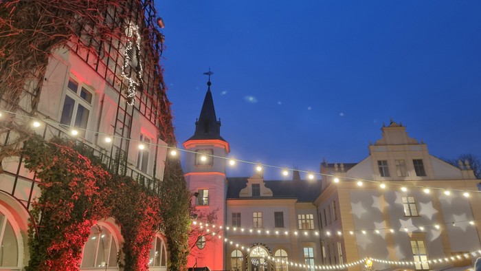 In der Abenddämmerung versammeln sich Menschenmengen auf einem Weihnachtsmarkt im Freien vor geschmückten Gebäuden mit Lichterketten.