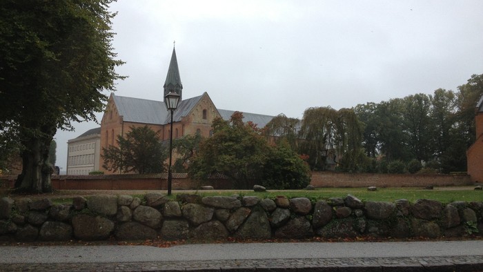 En charmerende murstenskirke med et højt spir står ved siden af et stengærdet græsareal og vejtræer.