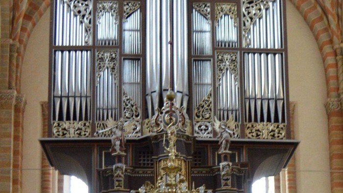Komplekst historisk pibeorgel med udsmykkede træskærerier og detaljer i guld fra en lysekrone i en stor kirke.