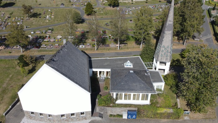 Ein weißes Gebäude mit schwarzem Dach neben einer Kirche mit hohem Turm und Parkplatz.