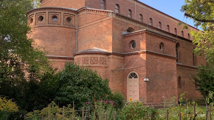Ein großes, rotes Backsteingebäude mit Rundbogenfenstern und Tür, umgeben von üppigem Grün und gelben Blumen.
