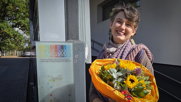 alttext bitte Eine lächelnde Frau mit kurzen braunen Haaren und einem großen Strauß Sonnenblumen steht im Sonnenschein vor dem Steinheimer Familien- und Generationenzentrum. Neben ihr ist das Eingangsschild der Einrichtung zu sehen.