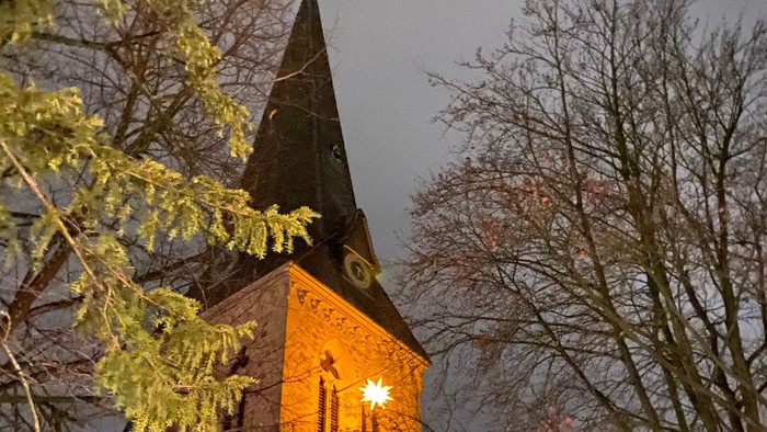 Ein historischer Kirchturm, beleuchtet in der Abenddämmerung, gerahmt von kahlen Winterbäumen.