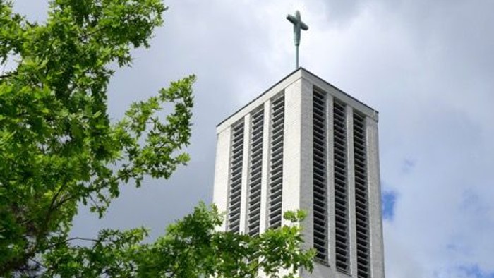 Moderner Kirchturm mit Kreuz, eingerahmt von üppigem grünem Laub unter einem bewölkten Himmel.