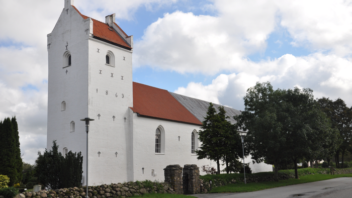 Et hvidt kirkebygning med rødt tag og tårn, omgivet af træer og en stenmur.
