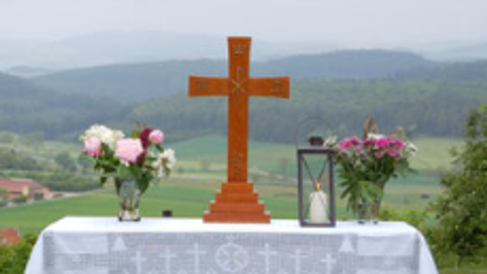 Das Foto zeigt einen aufgebauten Altar auf einer Wiese. Der Tisch trägt ein Kreuz, 2 Blumensträuße links und rechts und eine Kerze.